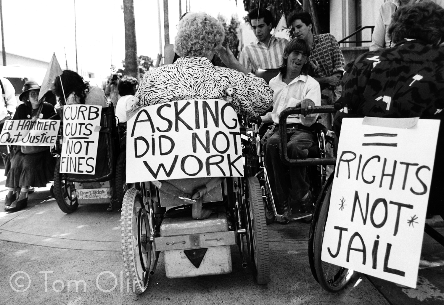 Black and white photograph of several disability activists in wheelchairs. They are holding signs that say I'd hammer out justice, curb cuts not fines, asking did not work, and rights not jail.