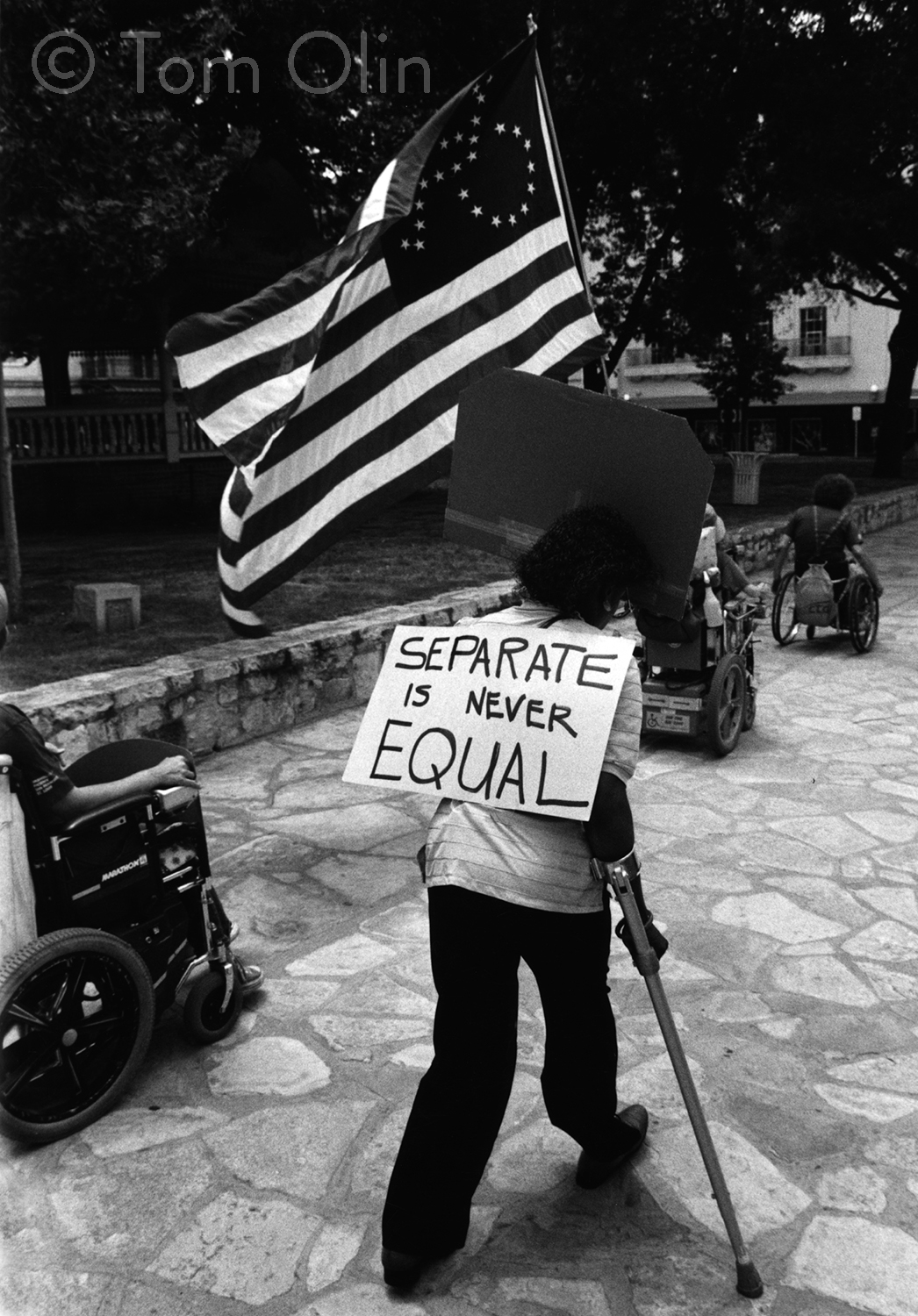 Black and white photograph of several disability rights activists marching on a cobblestone sidewalk. In the foreground, an activist his using a cane and holding a large American flag. Where the fifty stars usually are, there's an outline of the accessibility symbol made of stars. The activist is holding a sign that says separate is never equal. In the background are several people in wheelchairs.