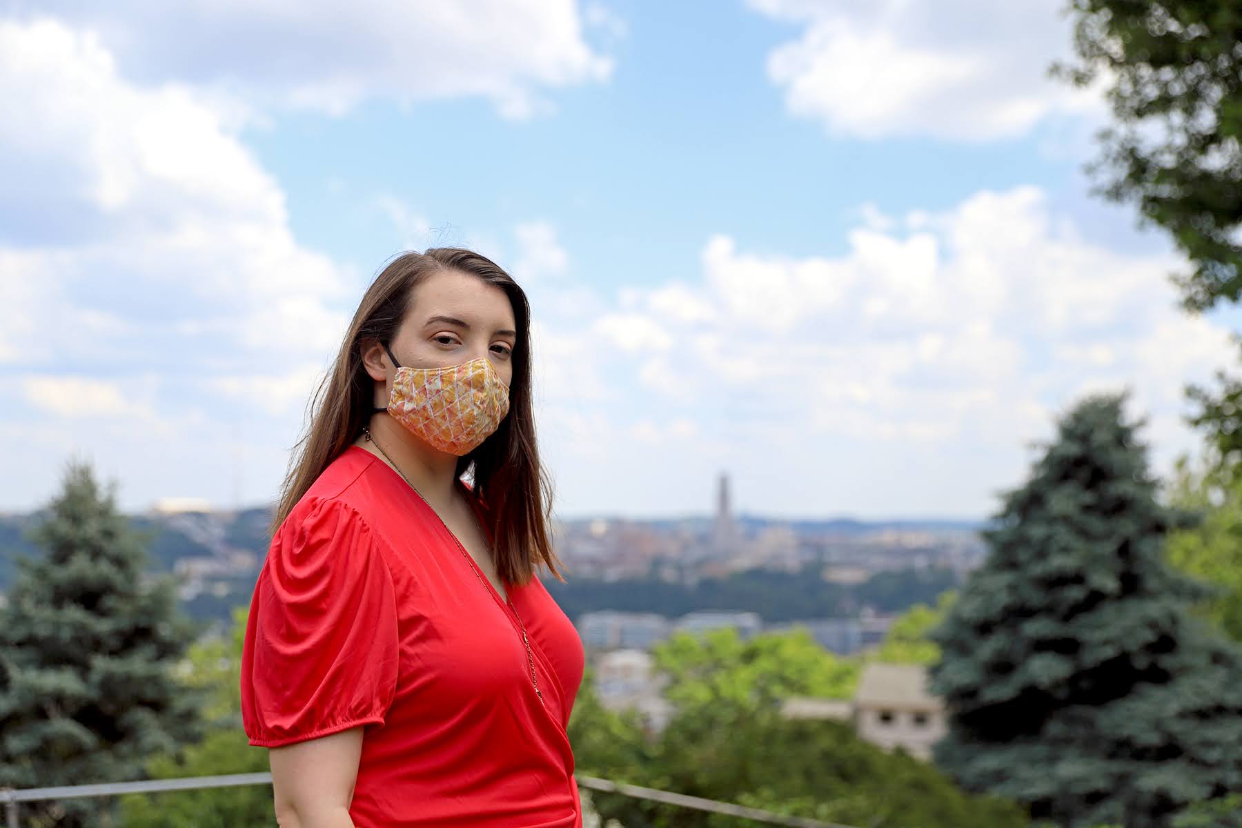 Photograph of essay author Jessica Benham standing outside with Downtown Pittsburgh in the background. She a white woman wearing a yellow mask and a bright pink shirt.