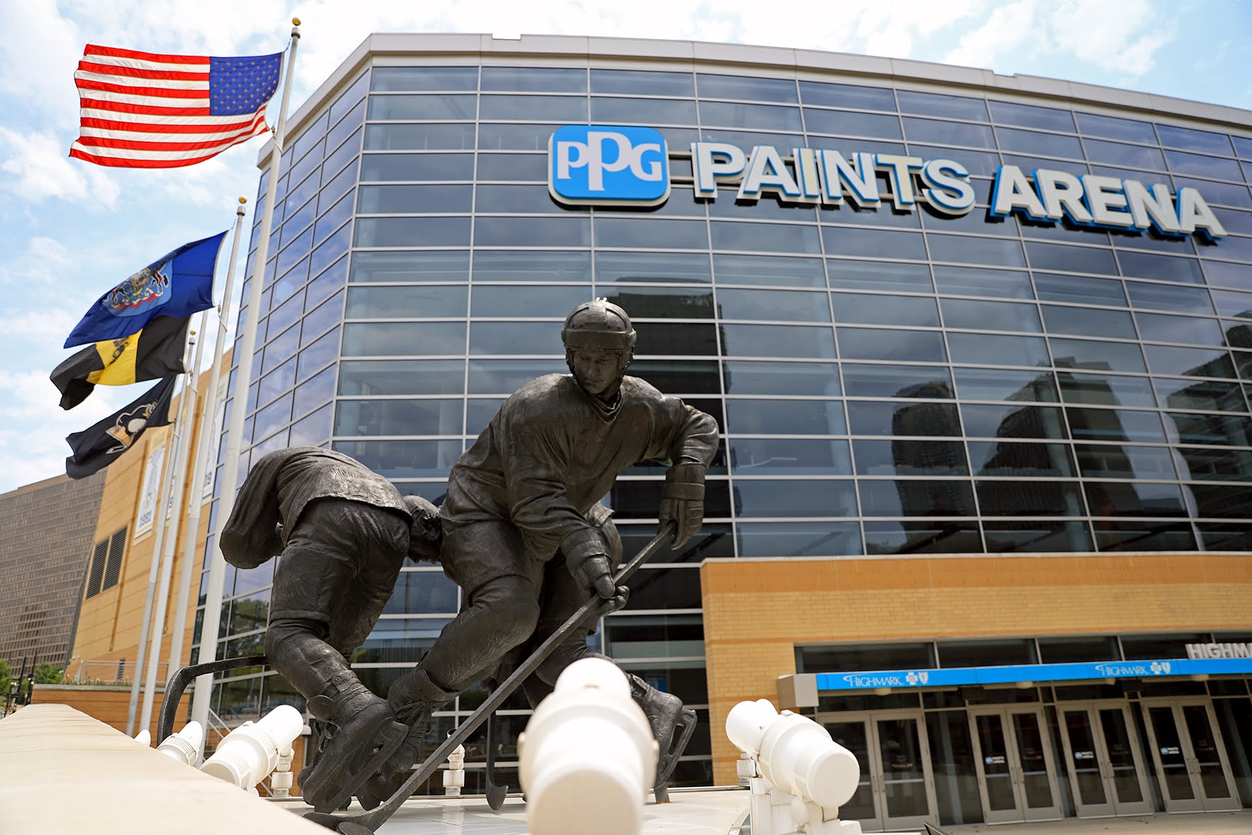 Photograph of the outside of the PPG Paints Arena, a large building made mostly of glass. In the foreground is a metal statue of two hockey players. In the background are the American, Pennsylvania, Pittsburgh, and Penguins flags.