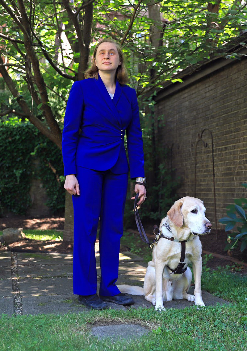 Photograph of essay author Catherine Getchell, a blind woman wearing a blue suit. She is standing in her backyard with her guide dog.
