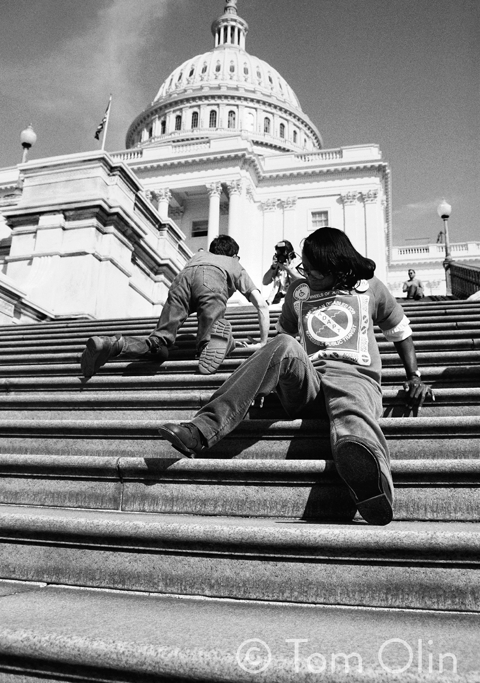 Black and white photograph of two disability rights activists. They are climbing the 83 steps up capitol hill. The photograph is shot from a low angle. In front, an activist is sitting and using their hands to push themself up the stairs backwards. In the background, another activist is crawling on his hands and knees.