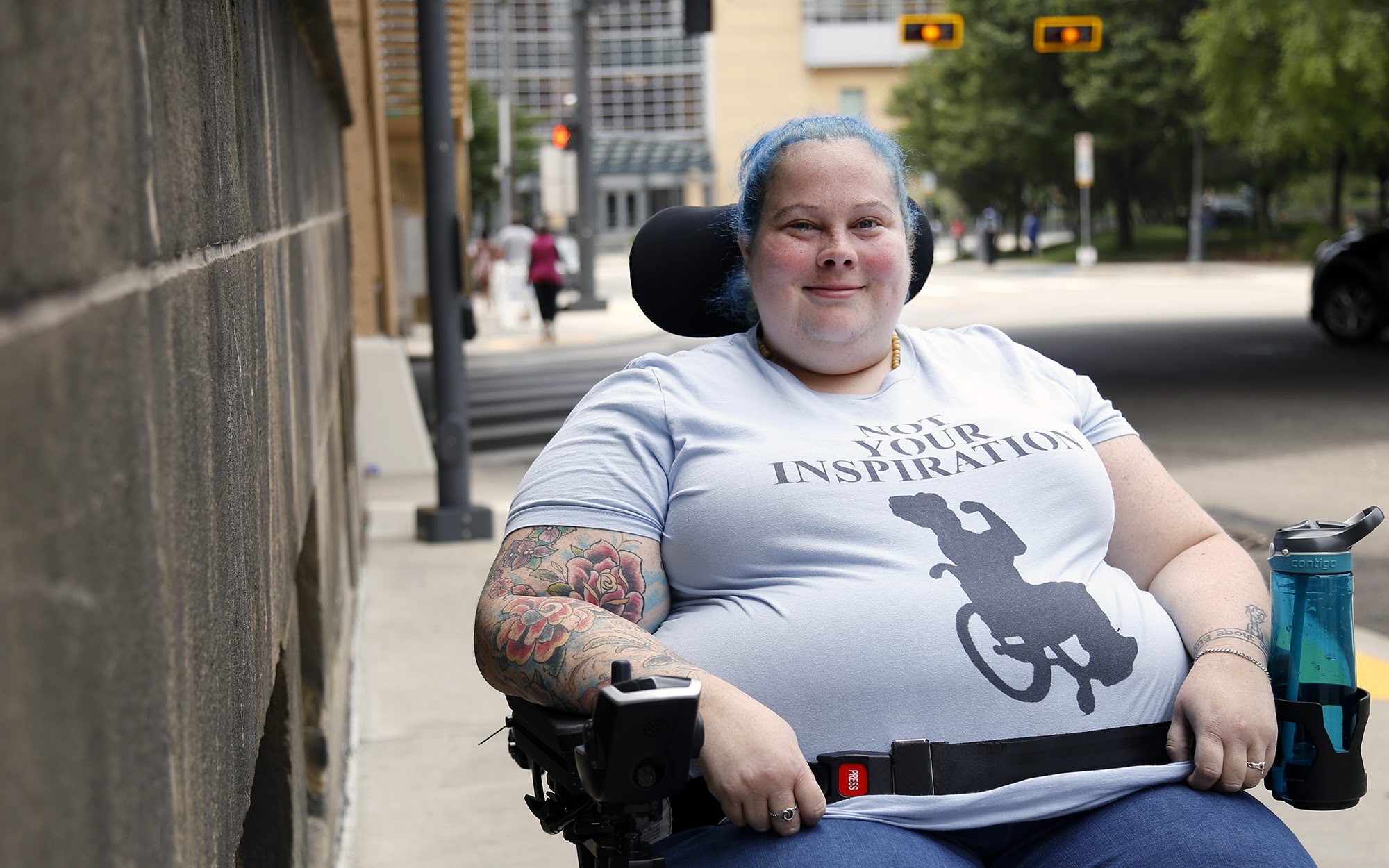 Photograph of Alisa Grisham outside on the sidewalk with the street behind her. She is in her wheelchair and wearing a T-shirt that says “not your inspiration” and below it is a silhouette of a person in a wheelchair.