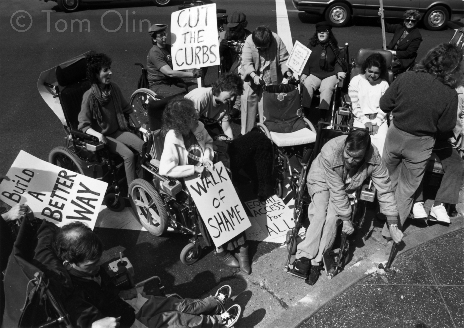 A black and white photo of a group of protestors in wheelchairs. One is using a sledgehammer to break up the curb. Several protestors hold signs saying “cut the curbs”; “build a better way”; and “walk of shame.”