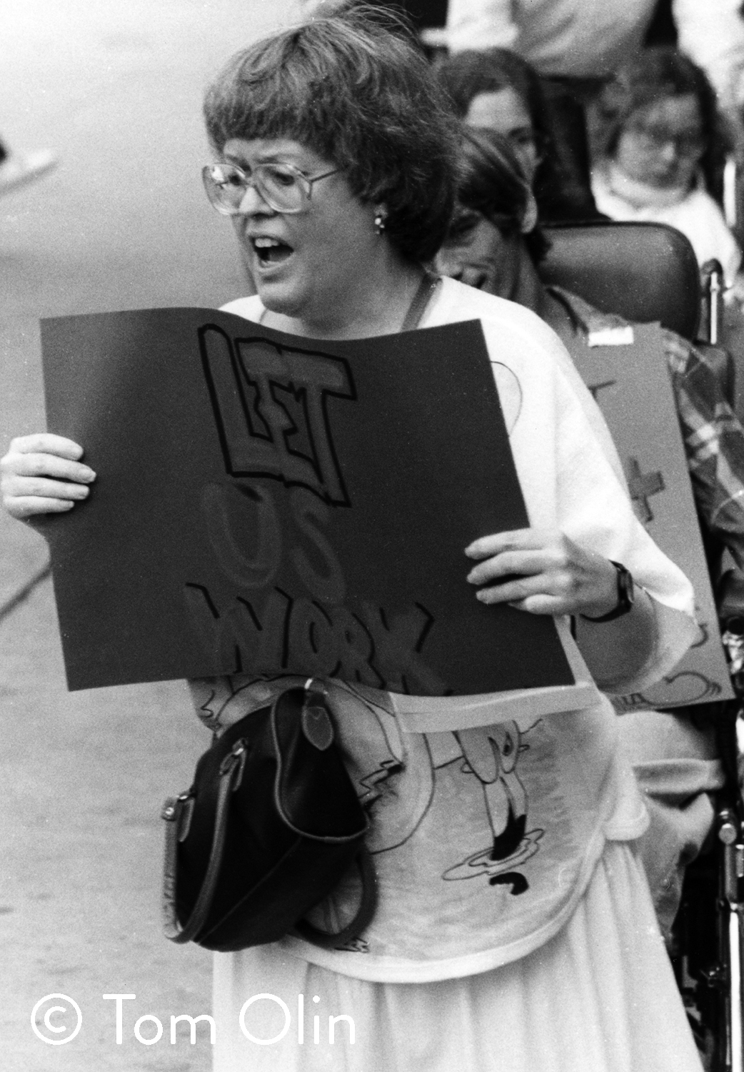 Black and white photograph of a woman protesting. She’s holding a handmade sign that reads “Let us work”