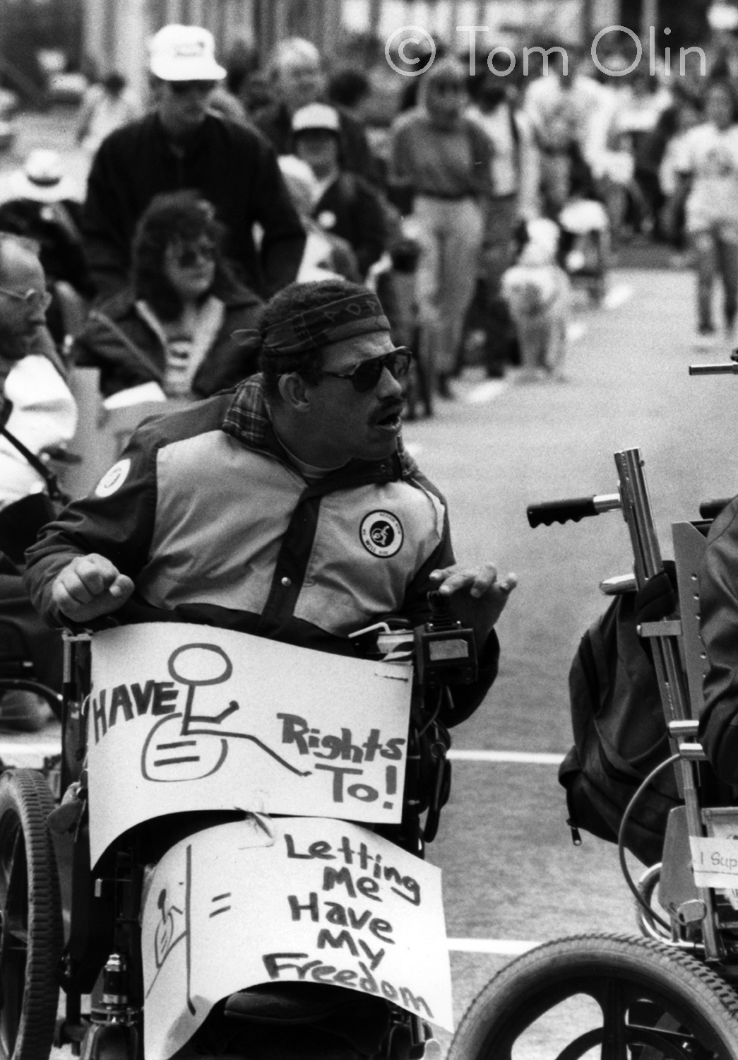 Black and white photograph of a crowd of disability rights activists. Many are in wheelchairs. In front, a Black man in a wheelchair is holding two signs. Both have drawings of wheelchairs on them. One says I have rights too! and the other says let me have my freedom.