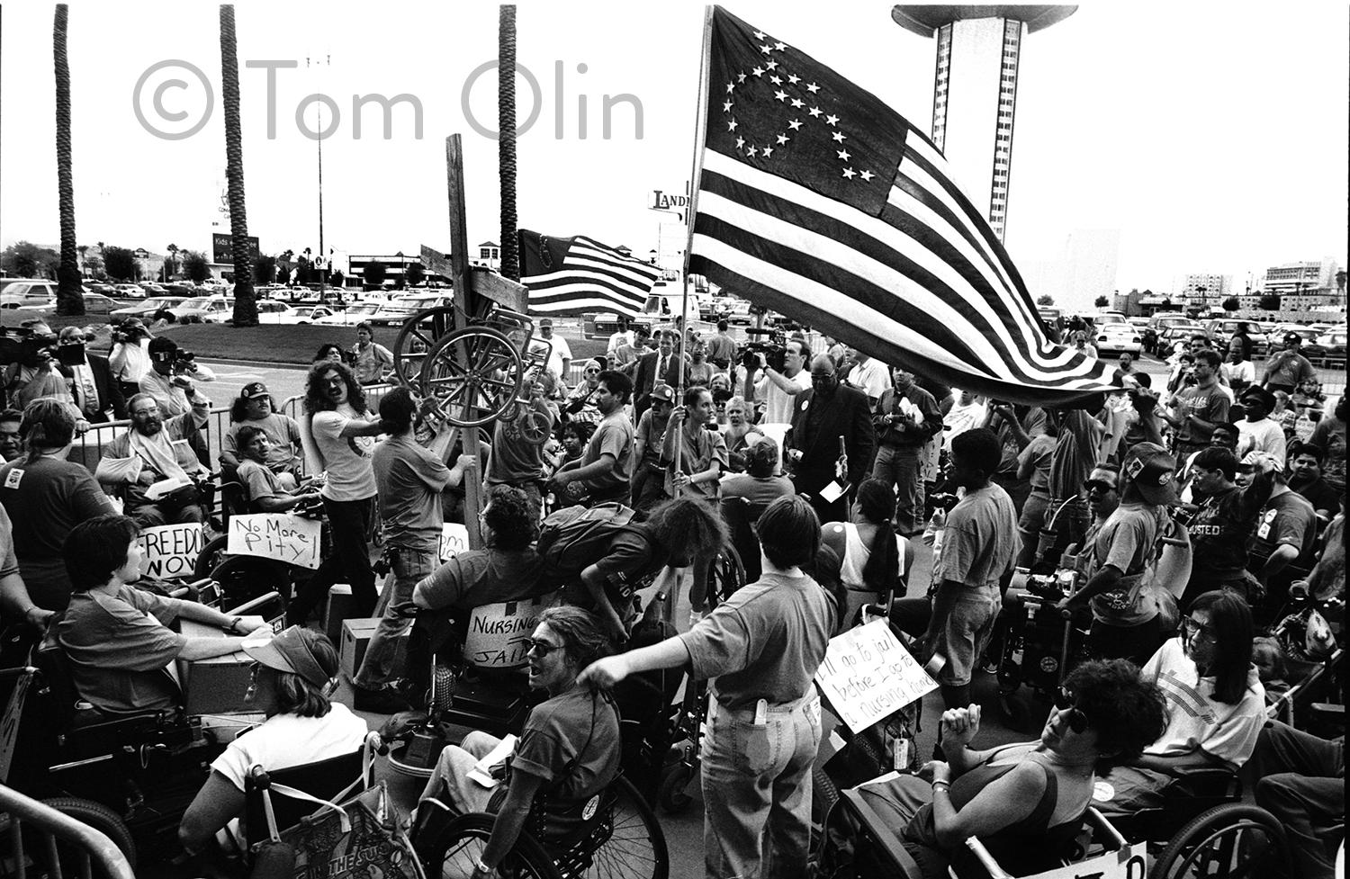 Black and white photo of a large group of activists. One is holding a large American flag. Where the fifty stars usually are, there's an outline of the accessibility symbol made of stars.