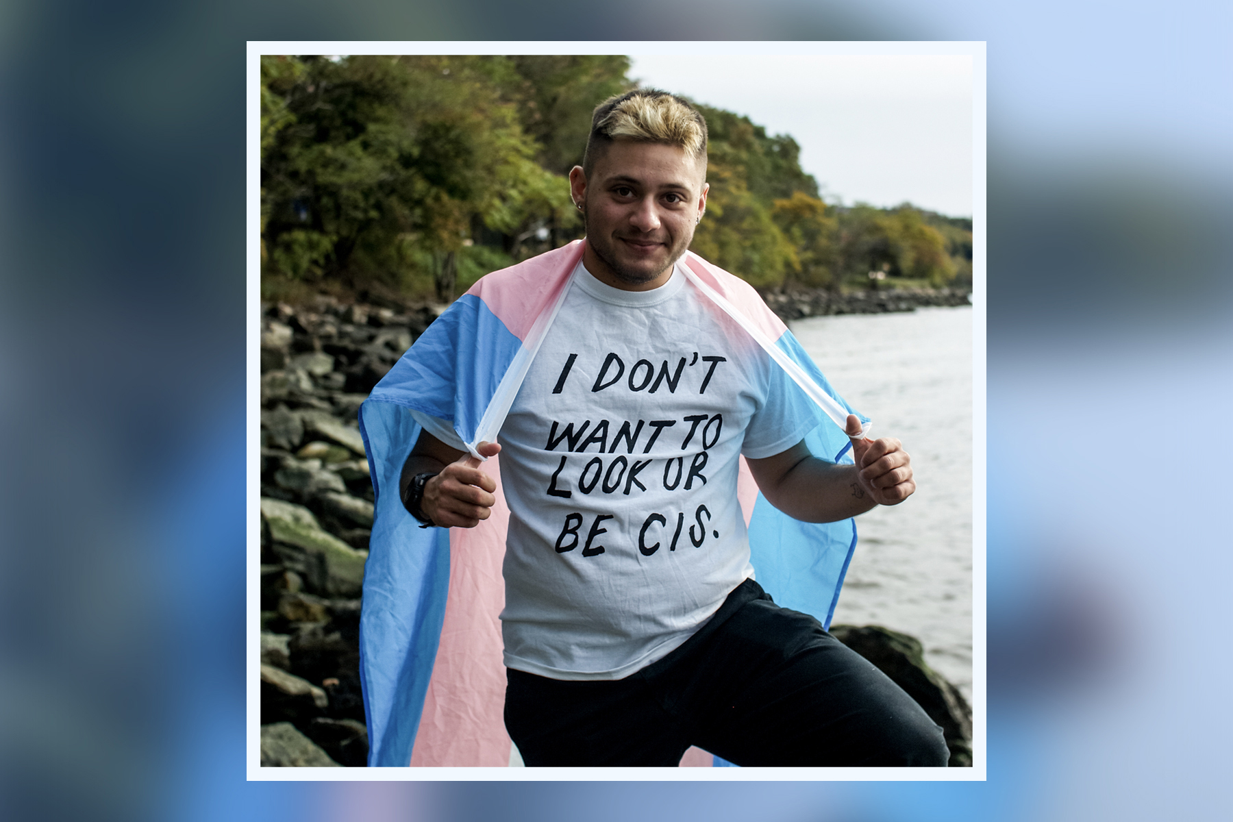 Photograph of essay author Dylan Kapit. They are standing outside looking straight at the camera. They're wearing a transgender pride flag around their shoulders and a shirt that says I don't want to look or be cis.