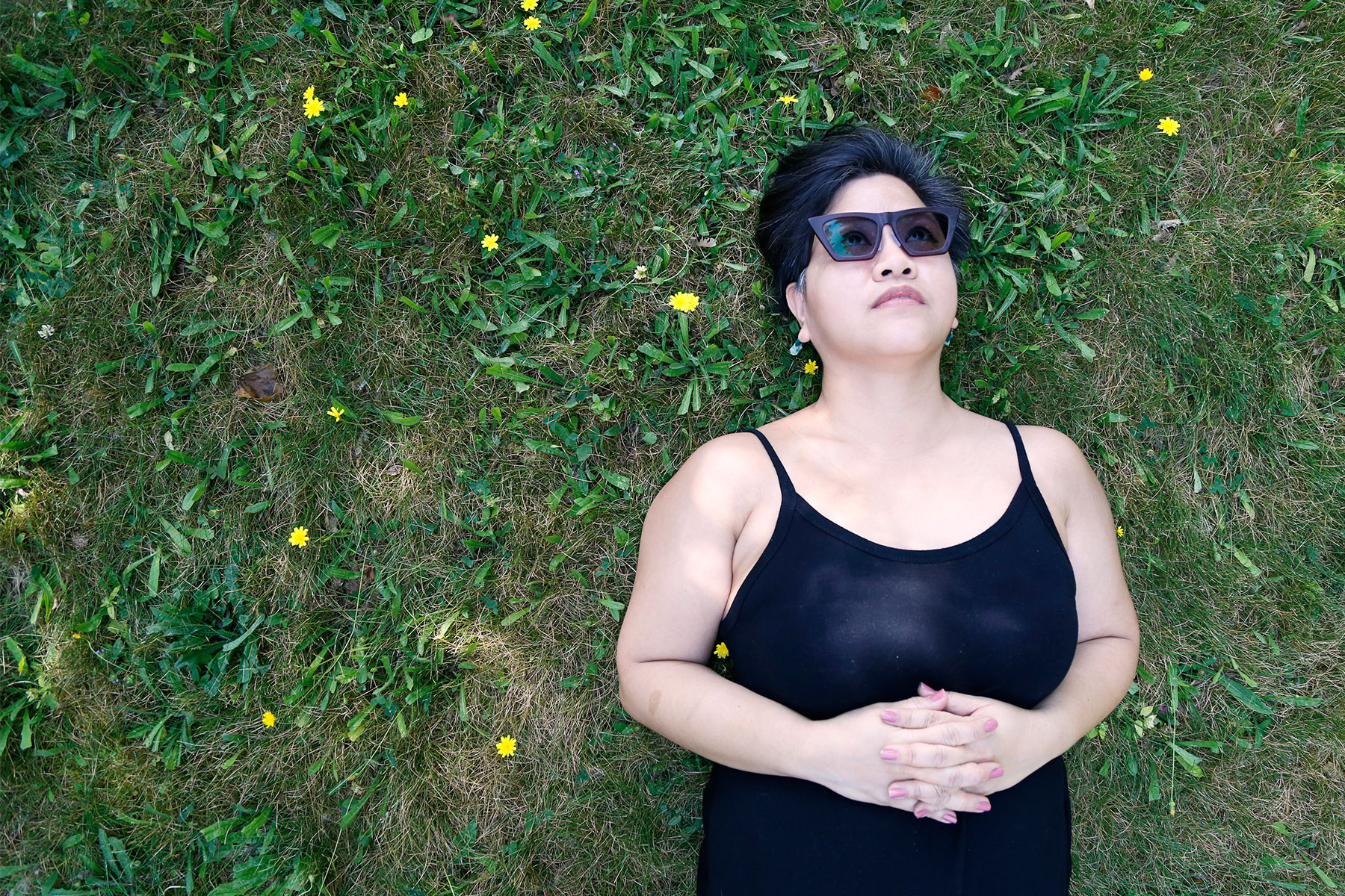 Photograph of essay author Fran Flaherty, a Filipino woman wearing sunglasses and a black jumpsuit. She's lying in the grass with small yellow wildflowers around her.