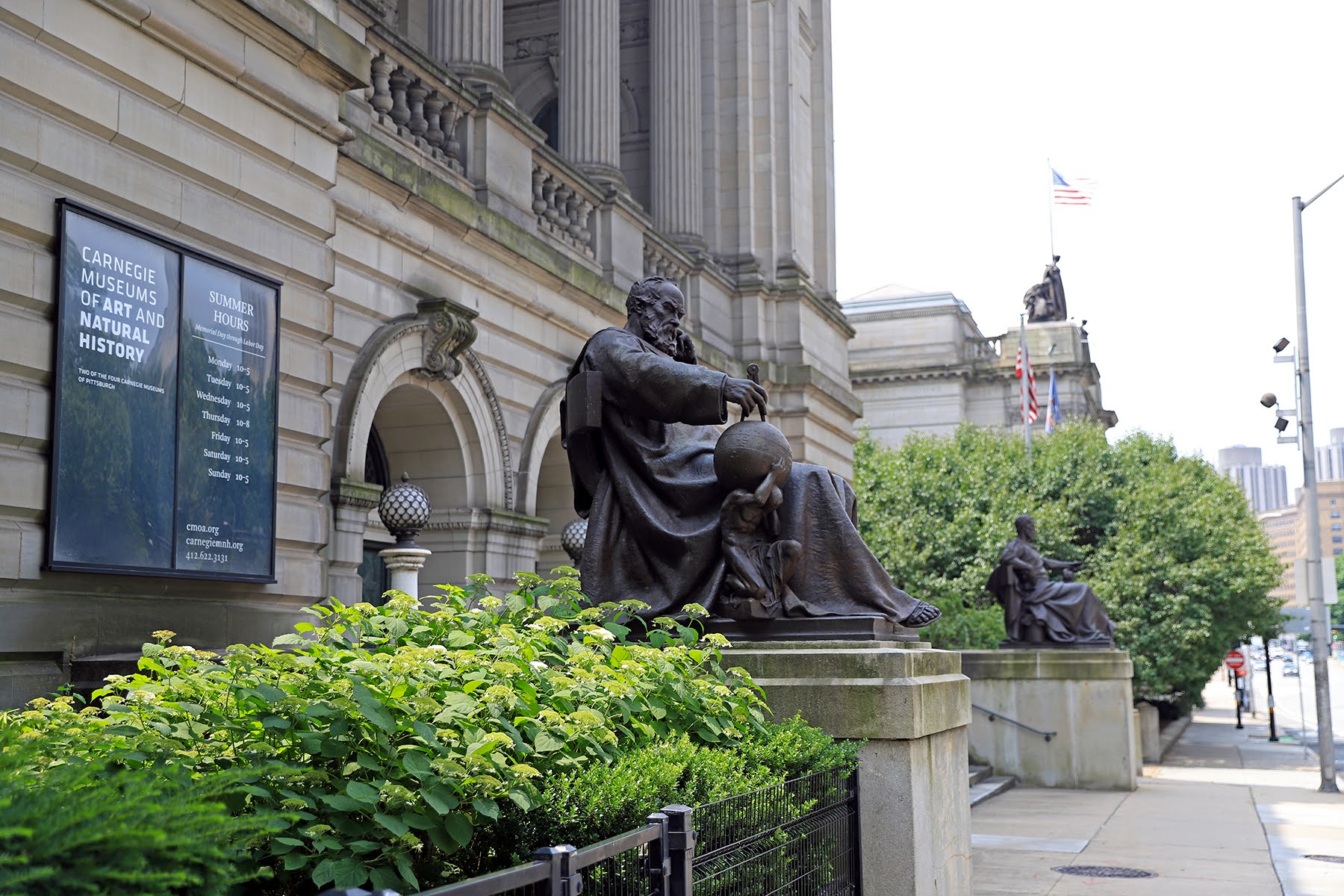 Photograph of the outside of the Carnegie Museums of Art and Natural History. It’s a big, classical style building with two metal statues of scientific leaders out front. Though this entrance has stairs, the museum also has an accessible entrance.