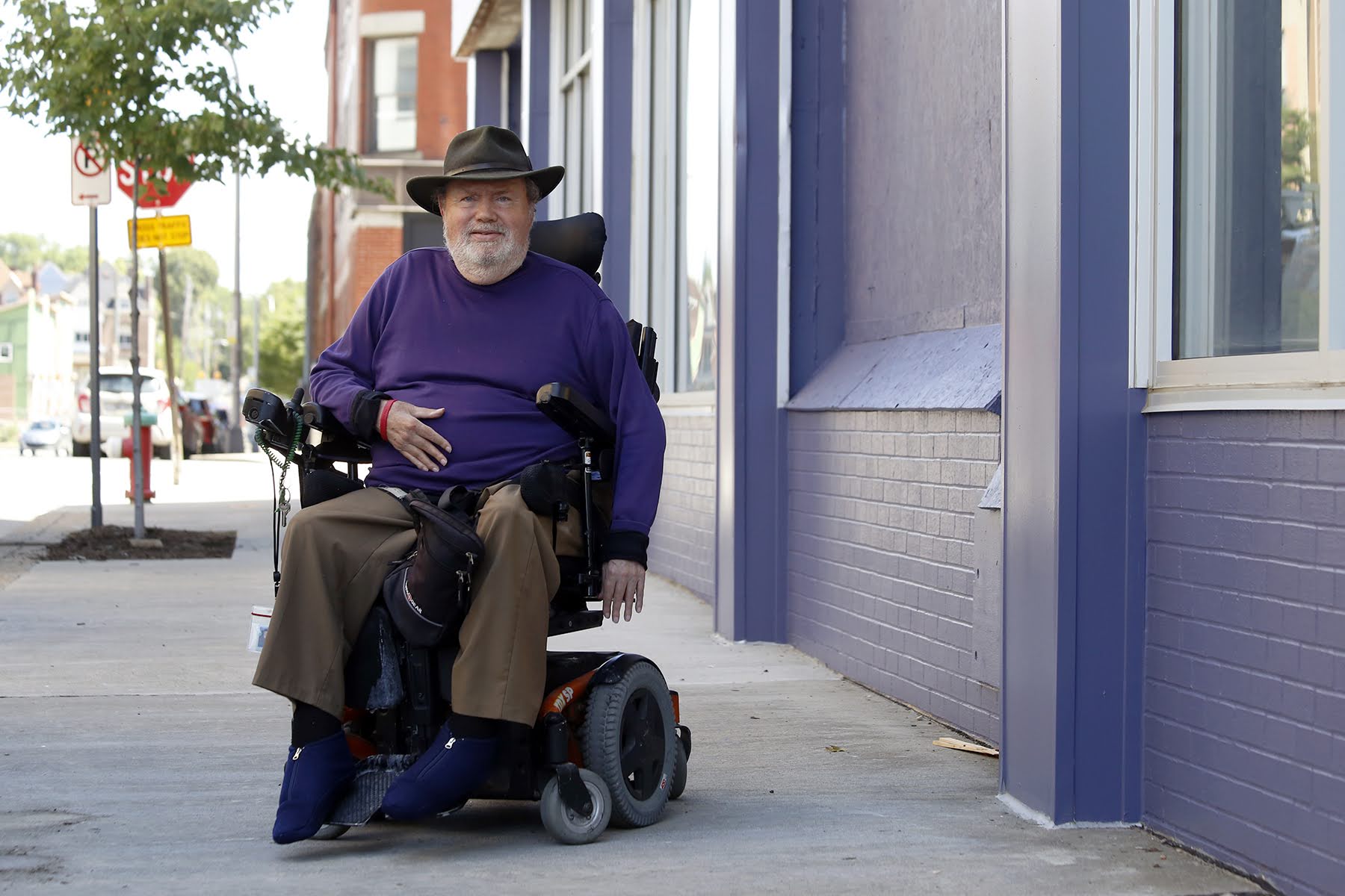Photograph of essay author John Tague, a white man in sitting in a power wheelchair. He's on the sidewalk facing the camera, wearing a purple sweater and wide hat.