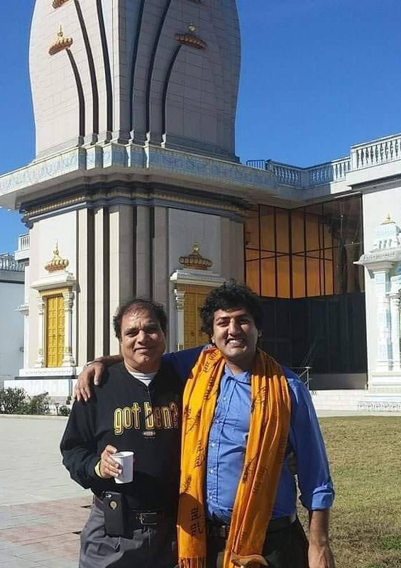 Photograph of two Brown men stand with their arms around each other in front of a Hindu temple. Both are facing the camera. Puneet stands on the right with a big smile on his face.