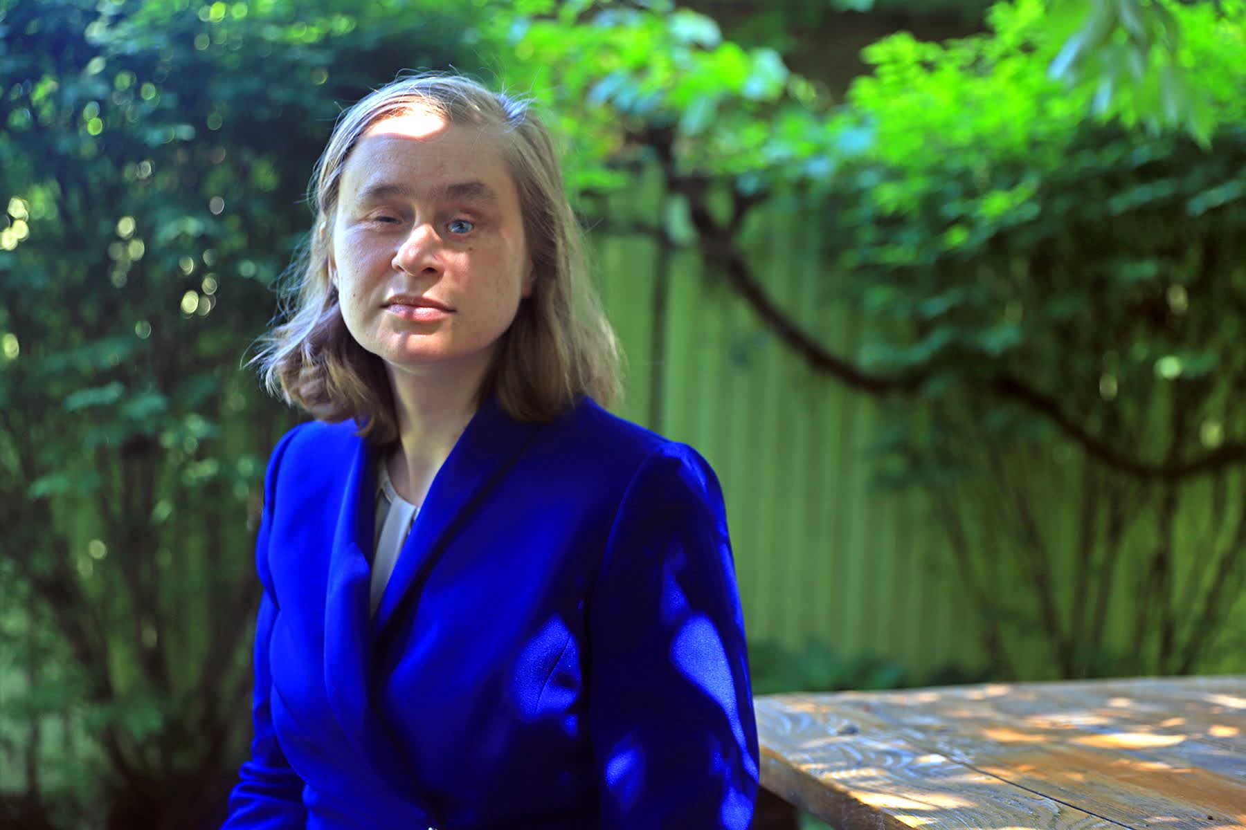 Close up photograph of essay author Catherine Getchell, a blind woman wearing a blue blazer. She’s sitting at a wooden picnic table in her backyard. There’s greenery in the background.