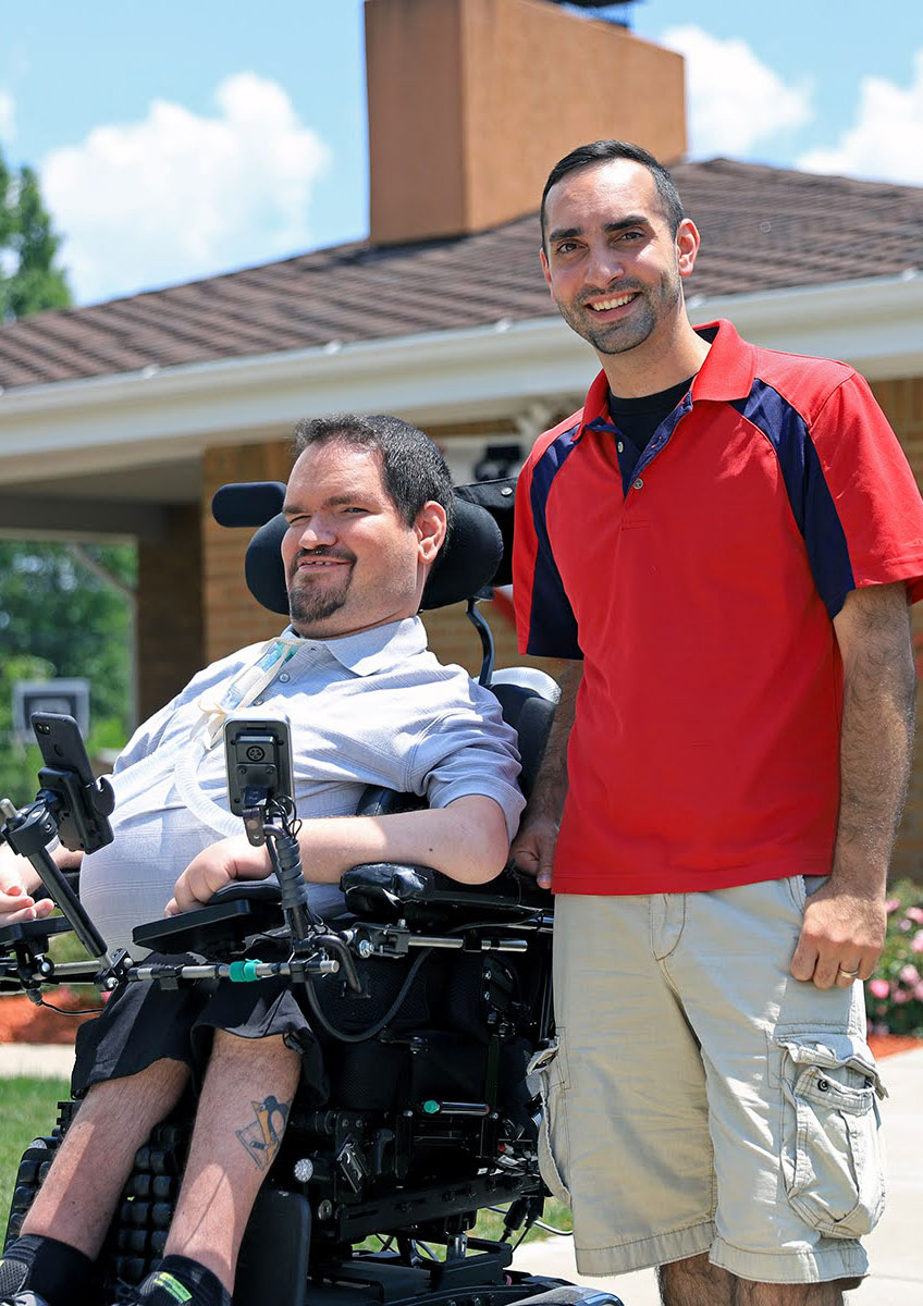 Photograph of brothers Nick and Eric Sinagra standing outside Nick's house. They're both smiling at the camera. Nick is sitting in a power wheelchair, and Eric is standing beside him.