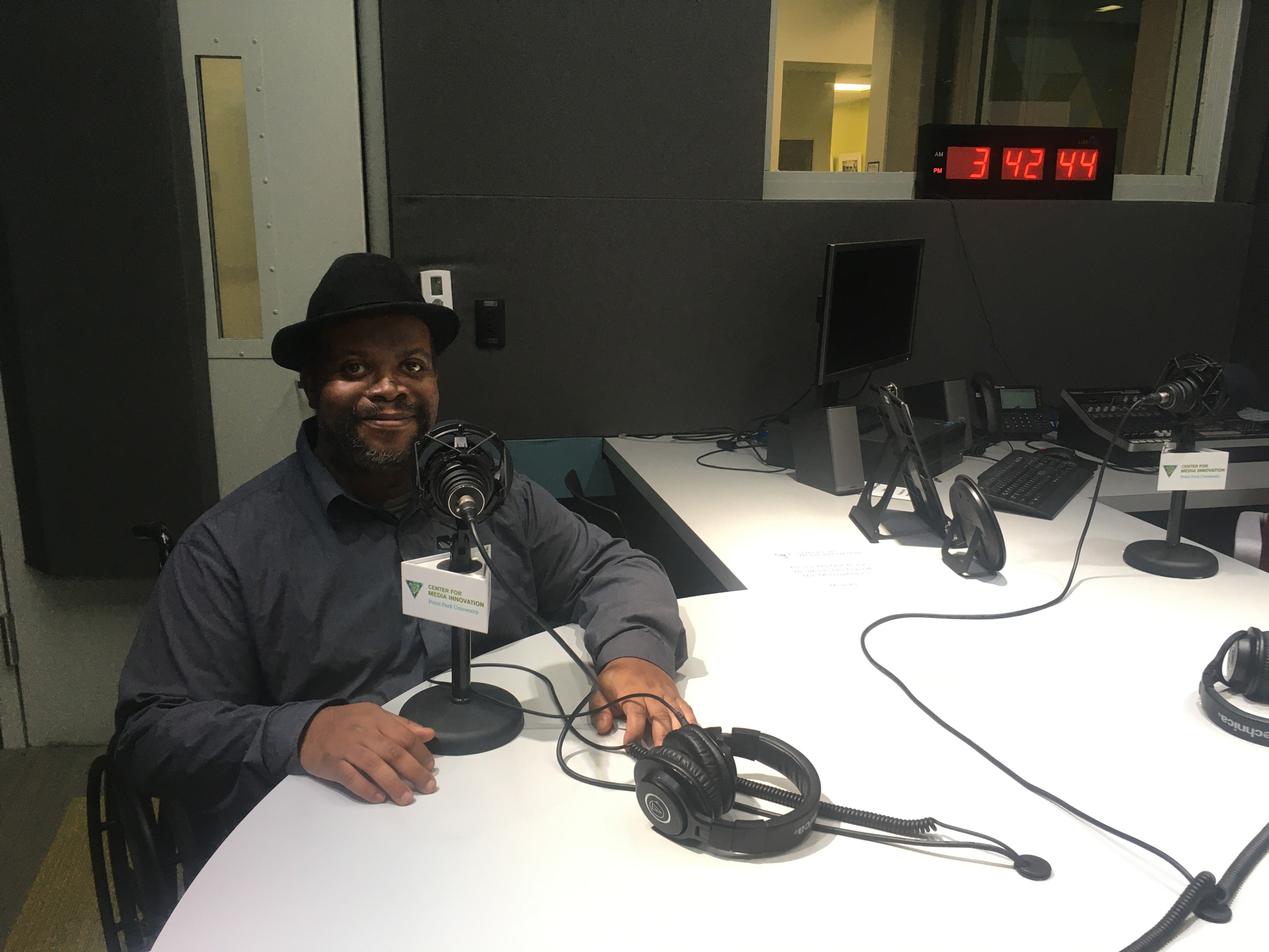Photograph of David Hale, a Black man wearing a fedora. He's sitting behind a microphone in a podcast studio.