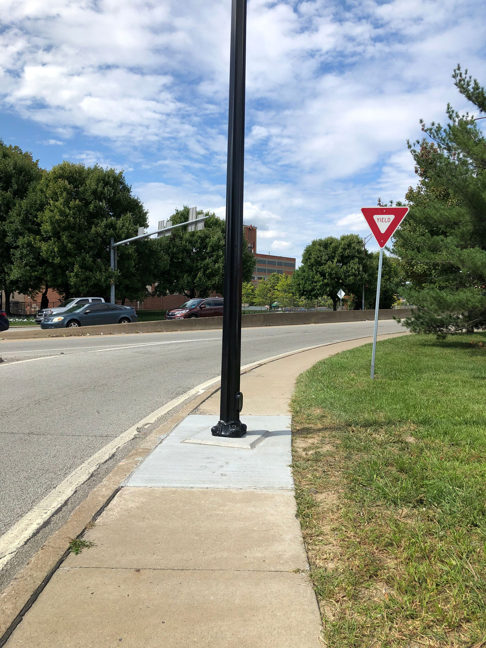 Photograph of a sidewalk. There’s a lamppost in the middle of the sidewalk, making it impossible for a wheelchair user to get past without going into the road or on the grass.