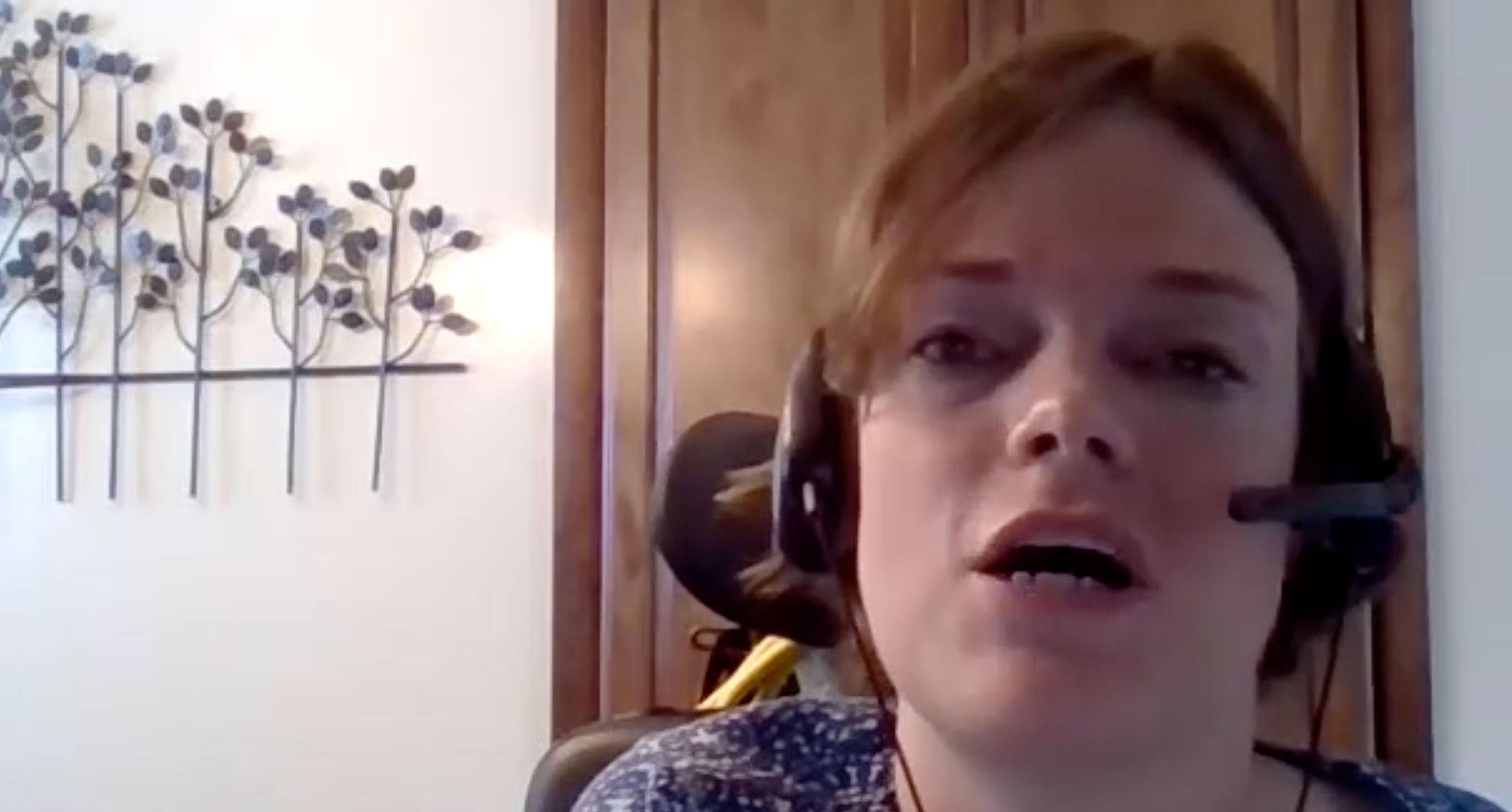Screen capture of a woman on video chat wearing a headset with microphone.