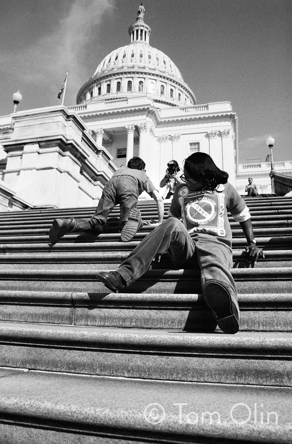 Black and white photograph of two disability rights activists. They are climbing the 83 steps up capitol hill. The photograph is shot from a low angle. In front, an activist is sitting and using their hands to push themself up the stairs backwards. In the background, another activist is crawling on his hands and knees.