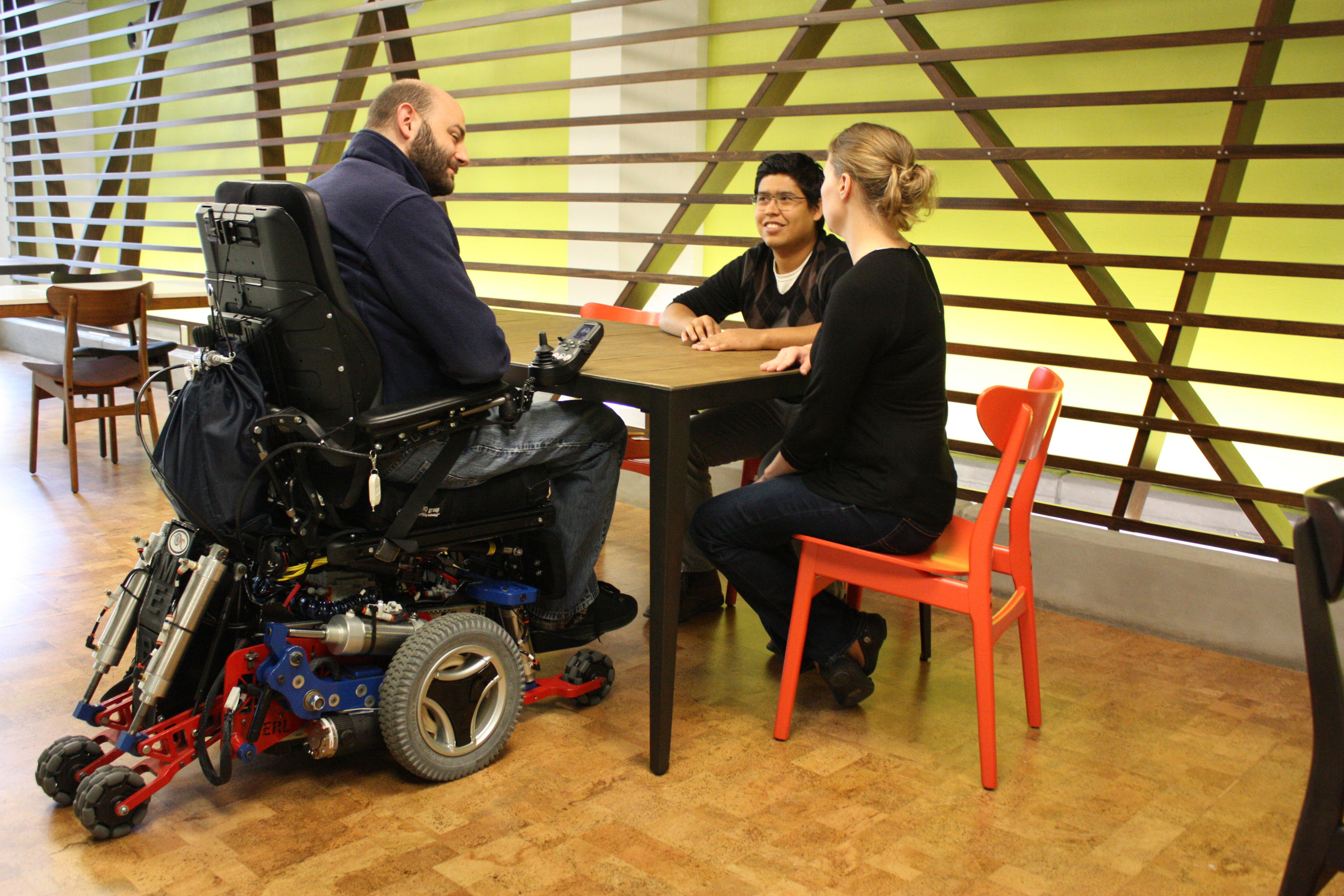 Photograph of three people sitting at a low table. In the foreground, a wheelchair user sitting in the MEBot is able to lower the height of his chair so he sits comfortably at the table.