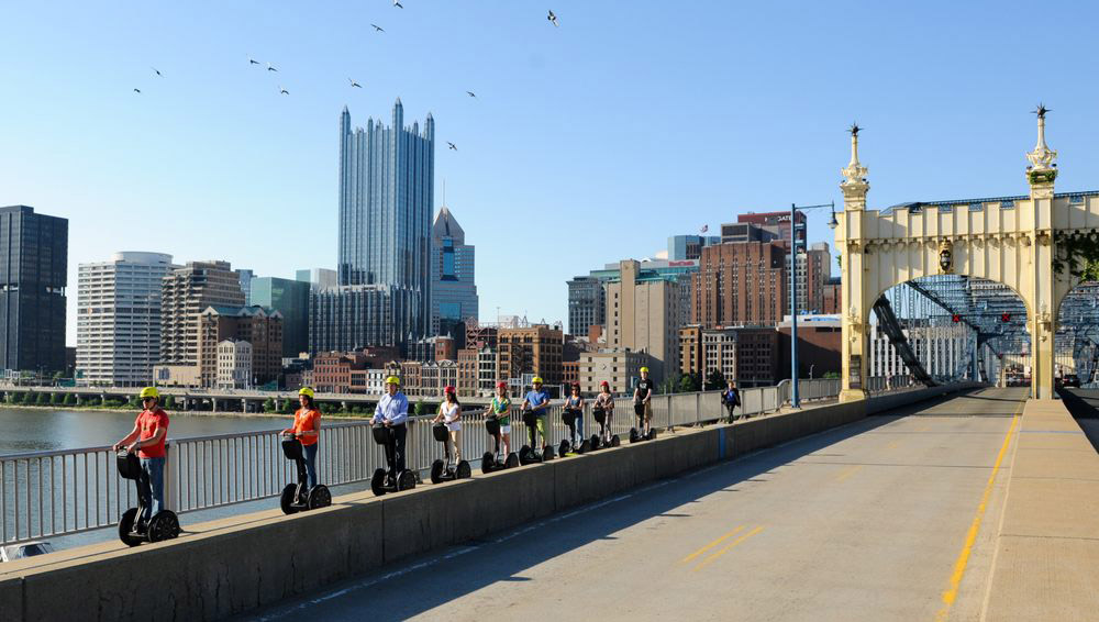 Wide shot photograph of a line of people riding segways. They're crossing a bridge in Pittsburgh. Behind them is Downtown Pittsburgh.