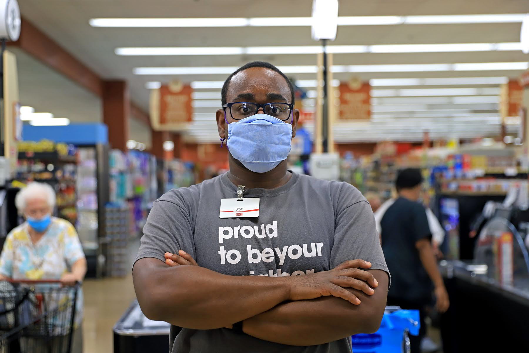 Photograph of essay author Joseph Vernon Smith. He is a Black man standing with his arms crossed looking at the camera. He wears a face mask and a Giant Eagle staff t-shirt that says proud to be your neighbor.