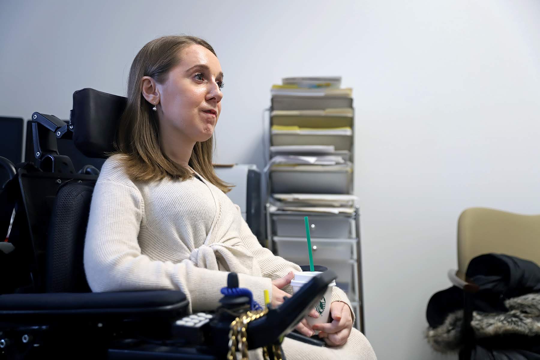 Photograph of Heather Tomko in her office. She is sitting in her wheelchair and holding a cup of coffee with a straw. Behind her are files, papers and folders.