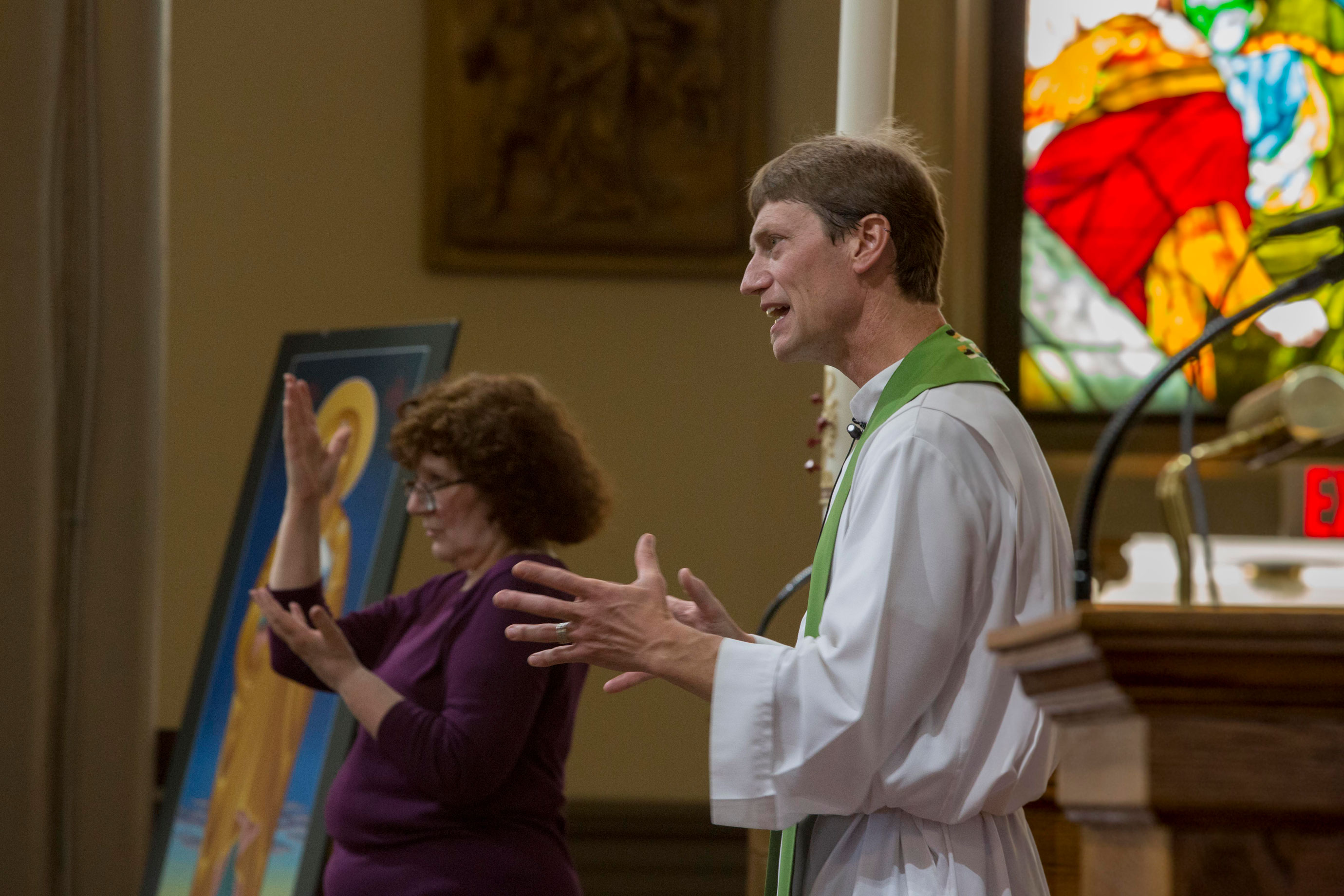 Photograph of a catholic priest delivering a sermon. He's inside a church in front of his congregation. In the background, a woman interprets his sermon in American Sign Language along with him.