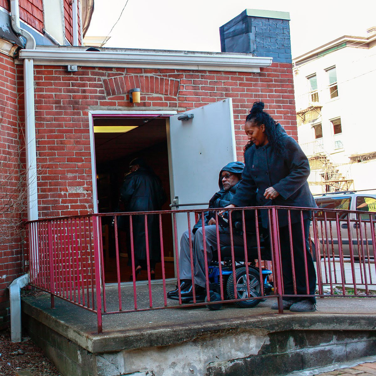 Photograph of an older Black man in motorized wheelchair going up a ramp into the back of a church. A younger Black woman walks beside him, helping him inside.